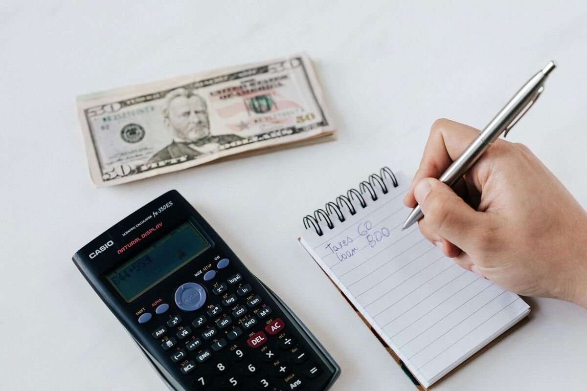 Hand writing loan and tax details in a notebook with calculator and dollar bills on a desk